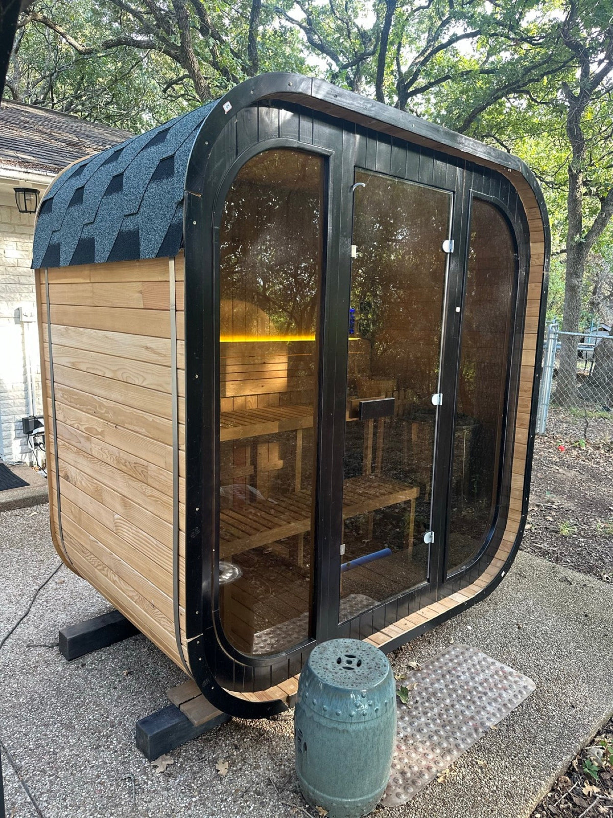 Medium 21 Cube outdoor sauna with full glass front, black frame and shingle roof in a backyard